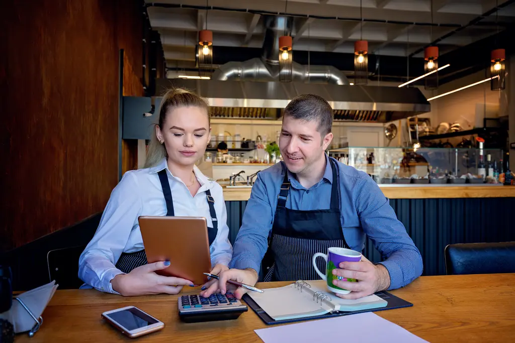 Two restaurant owners in aprons reviewing numbers on a tablet at a dining table