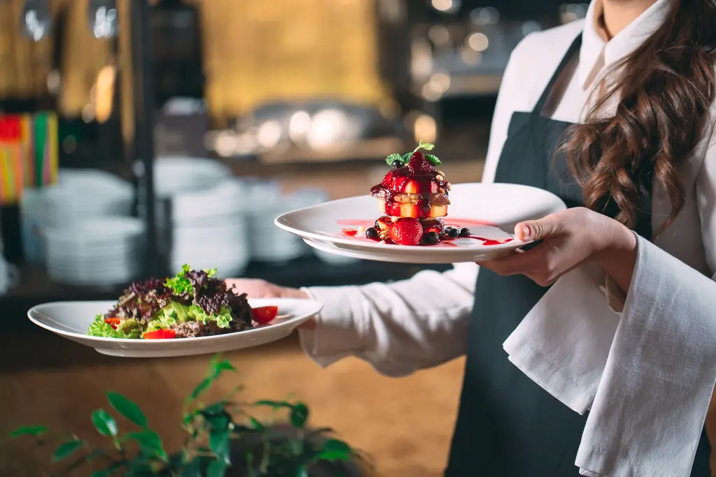 A server carrying plated dishes through a busy restaurant dining room